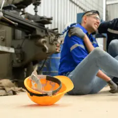 Robotnik po wypadku, kolega pomaga. Pomarańczowy kask leży obok. Sprawa dotyczy zadośćuczynienia a odszkodowania.