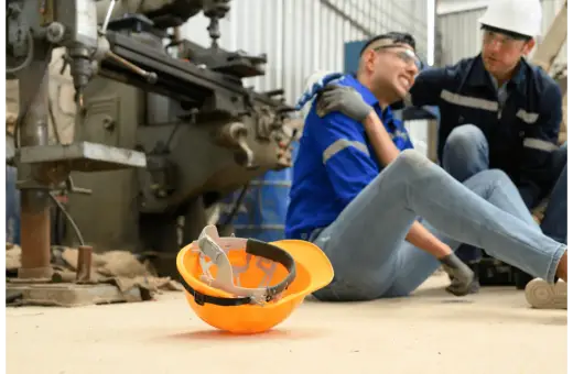 Robotnik po wypadku, kolega pomaga. Pomarańczowy kask leży obok. Sprawa dotyczy zadośćuczynienia a odszkodowania.