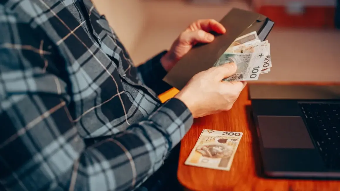 Osoba w kratę liczy pieniądze, przygotowując donos do urzędu skarbowego. Obok leży laptop i banknot 200 zł.